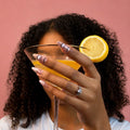 Evil Eye press-on nails, pink shimmer with black line art eyes, held around a martini glass with a lemon slice