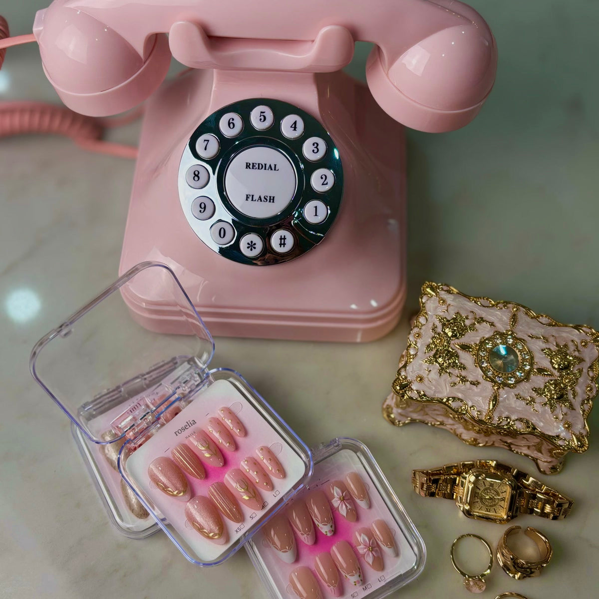 Gilded Tulip press-on nails, pink shimmer with gold tulip line art, in their case beside a pink rotary telephone and vintage jewellery