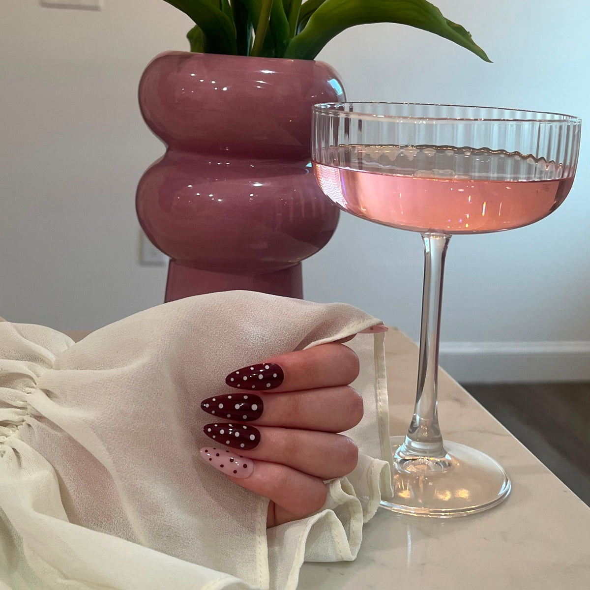 Merlot Dot press-on nails, deep burgundy with white polka dots, beside a glass of rose wine
